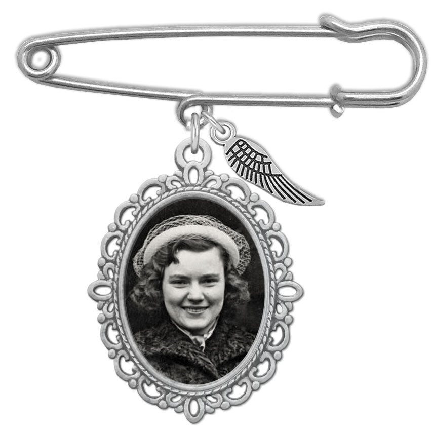 A silver wedding bouquet photo charm with safety pin style top featuring an oval filigree pendant with a black-and-white portrait photo in the frame and a small wing charm, shown on a plain white background.