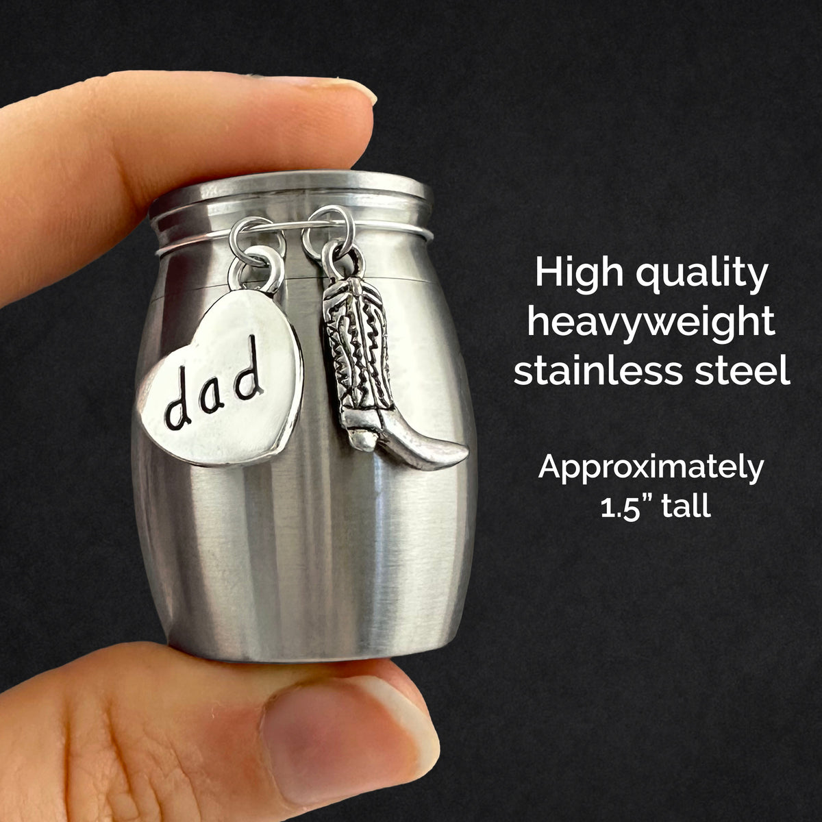 A hand holds a small stainless steel cremation urn shaped like a jar, with two dangling charms: a heart engraved with the word 'dad' and a cowboy boot. White text on the right reads 'High quality heavyweight stainless steel' and 'Approximately 1.5 inches tall.