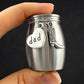 A hand holding a small brushed silver jar-shaped keepsake or cremation urn with two dangling charms: a heart charm stamped with the word "dad" and a small cowboy boot charm, against a dark background.