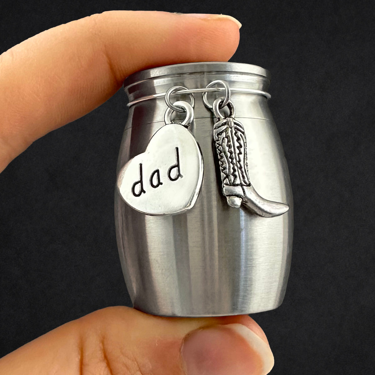 A hand holding a small brushed silver jar-shaped keepsake or cremation urn with two dangling charms: a heart charm stamped with the word "dad" and a small cowboy boot charm, against a dark background.