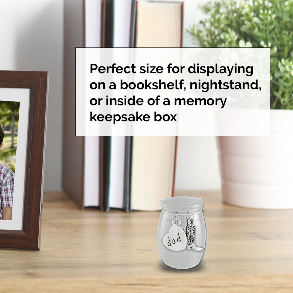 A small stainless steel urn with a lid and charm, labeled 'dad', displayed on a wooden surface with a blurred background of books, a plant, and a picture frame. A text box explains its use for bookshelves, nightstands, or memory keepsake boxes.