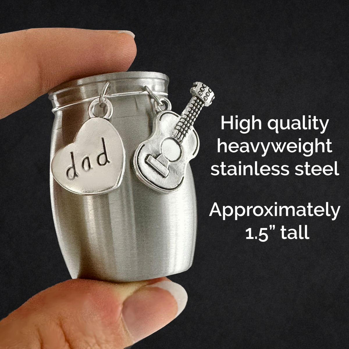 A hand holds a small brushed stainless steel urn ashes holder with a hanging heart charm stamped 'dad' and a guitar charm, against a dark background with text reading 'High quality heavyweight stainless steel' and 'Approximately 1.5" tall'.