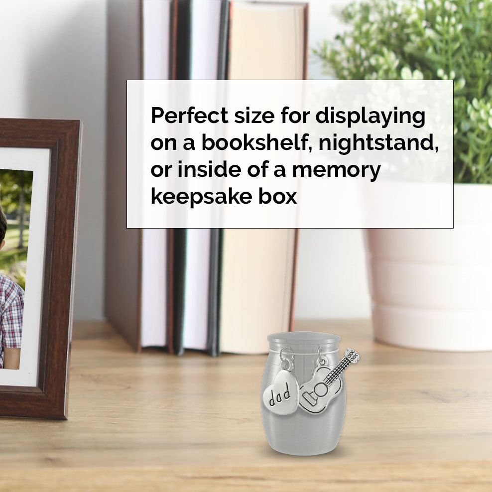 A small keepsake ashes holder urn with two silver charms, one shaped like a heart labeled 'dad' and the other shaped like a guitar, displayed on a wooden tabletop in a home setting. A text overlay explains the mini urns use for bookshelves, nightstands, or memory keepsake boxes.