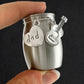 A small brushed silver metal jar-shaped keepsake or gift container held between fingers, with two hanging charms: a heart charm stamped with the word "dad" and a guitar-shaped charm.