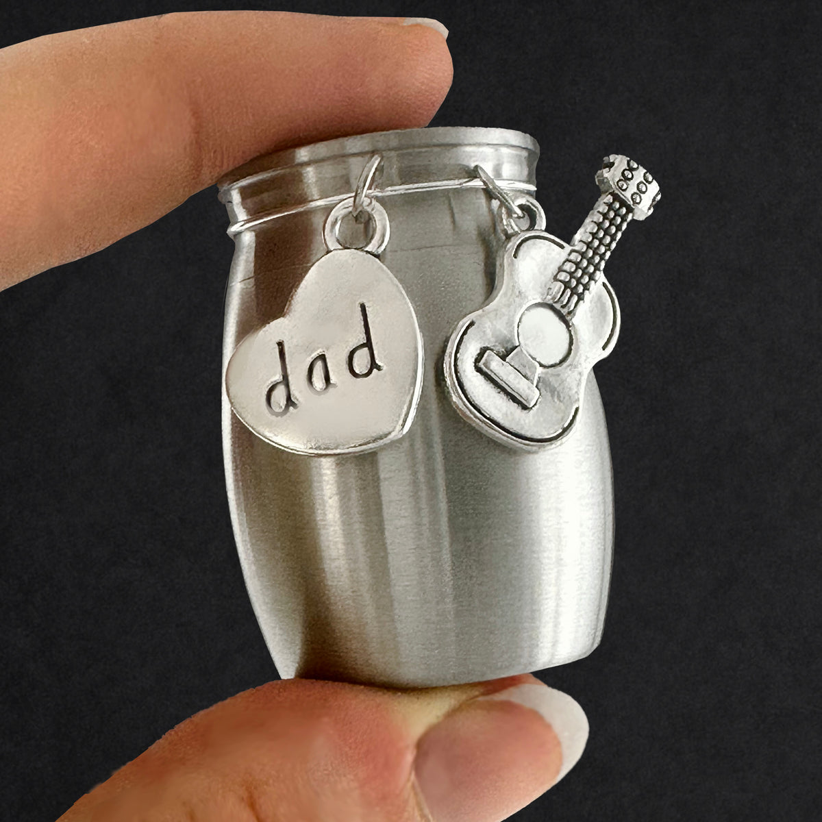 A small brushed silver metal jar-shaped keepsake or gift container held between fingers, with two hanging charms: a heart charm stamped with the word "dad" and a guitar-shaped charm.