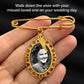Gold wedding bouquet photo charm pin with angel wing charm and ornate oval frame is held in a hand against a dark background. The pendant has an ornate filigree frame and a small wing detail, with a black-and-white portrait insert.