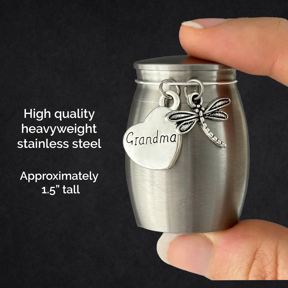 A hand holds a small brushed stainless steel keepsake urn, a heart-shaped charm engraved 'Grandma,' and a dragonfly charm. Text on the left reads 'High quality heavyweight stainless steel' and 'Approximately 1.5 inches tall'.