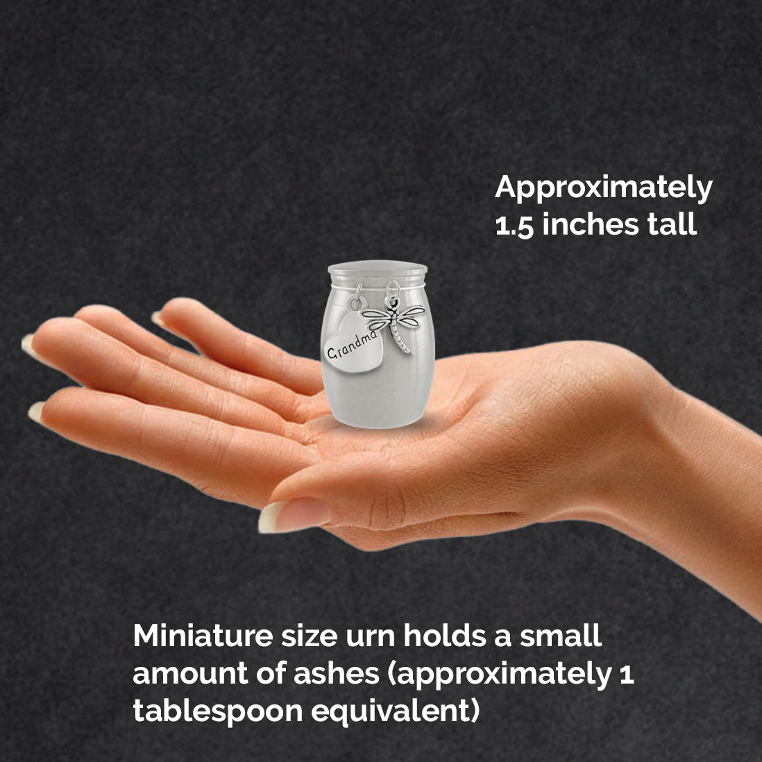 A hand holding a miniature stainless steel cremation ashes holder with  silver dragonfly charm and heart shaped charm that says 'grandma', set against a dark textured background with informational text about its size and ash capacity.