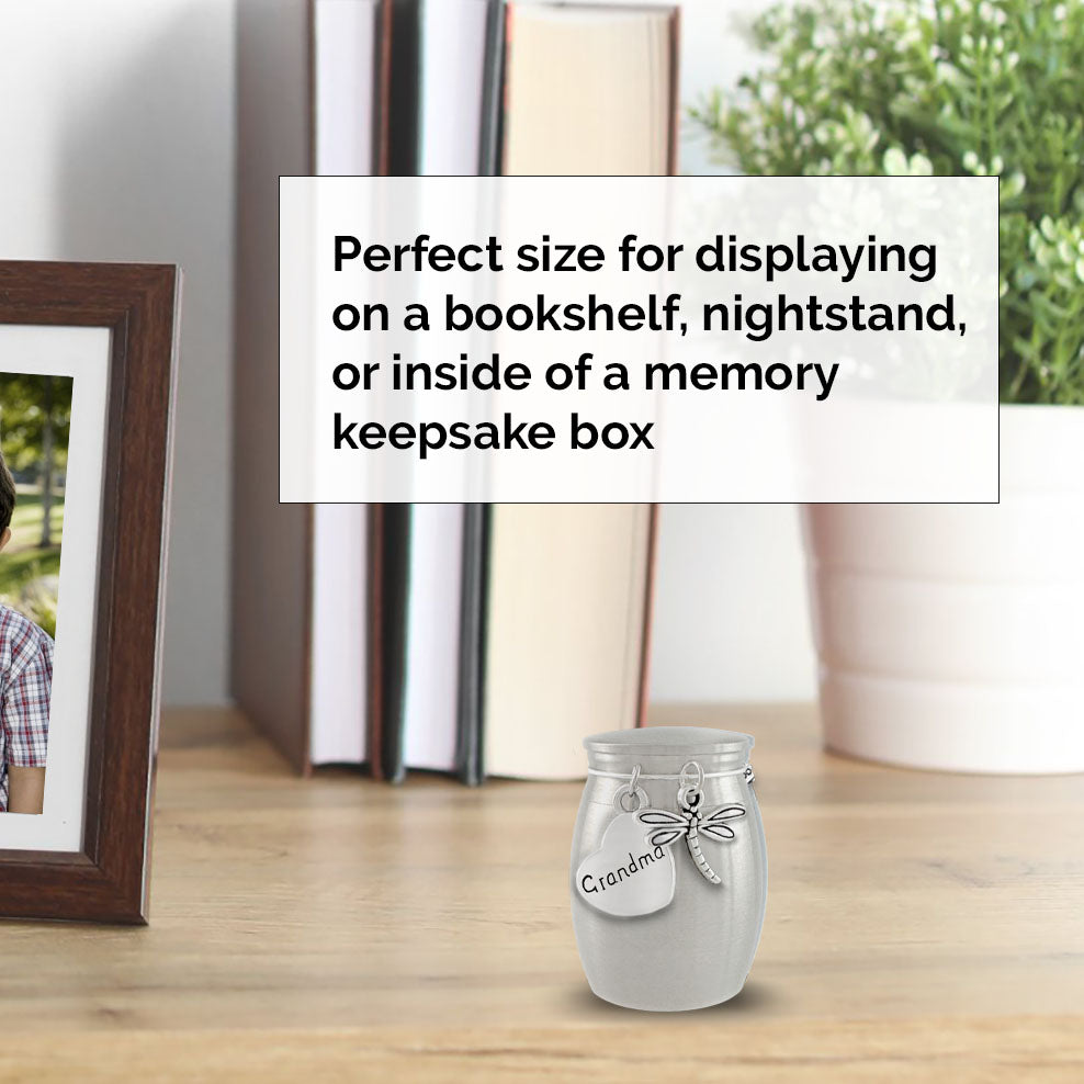A stainless steel miniature urn with dragonfly charm and a charm engraved 'Grandma' sits on a wooden table in a styled home setting with a bookshelf, books, a framed picture, and a potted plant. A text box explains its use for displaying on a bookshelf, nightstand, or inside a memory keepsake box.