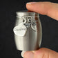 A small brushed-metal keepsake urn being held by a hand, with a heart-shaped charm engraved 'Grandma' and a dragonfly charm attached to the lid.