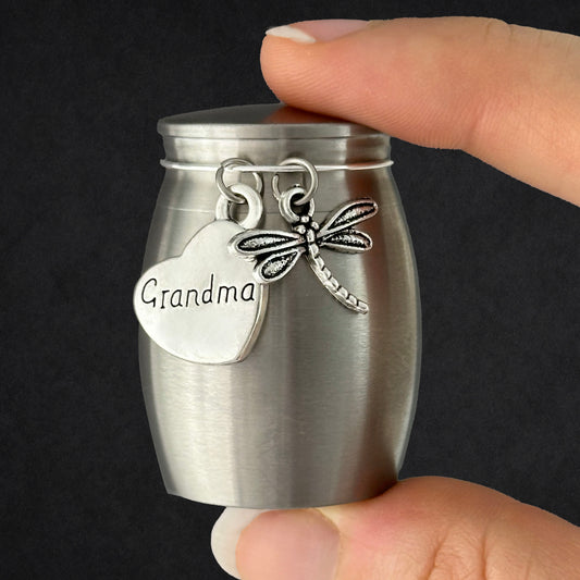 A small brushed-metal keepsake urn being held by a hand, with a heart-shaped charm engraved 'Grandma' and a dragonfly charm attached to the lid.