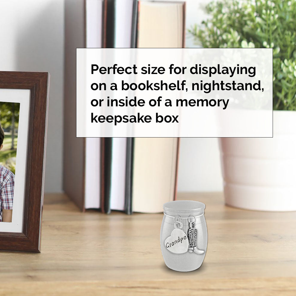 A small keepsake urn with a cowboy boot charm and a heart-shaped charm engraved with the text 'Grandpa' sits on a wooden tabletop. A framed photo, books, and a potted plant are blurred in the background, with a text box explaining use for bookshelves, nightstands, or memory keepsake boxes.
