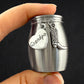 A hand holding a small brushed silver metal keepsake urn with hanging charms, including a heart-shaped tag stamped with the name 'Grandpa' and a cowboy boot charm, against a dark background.