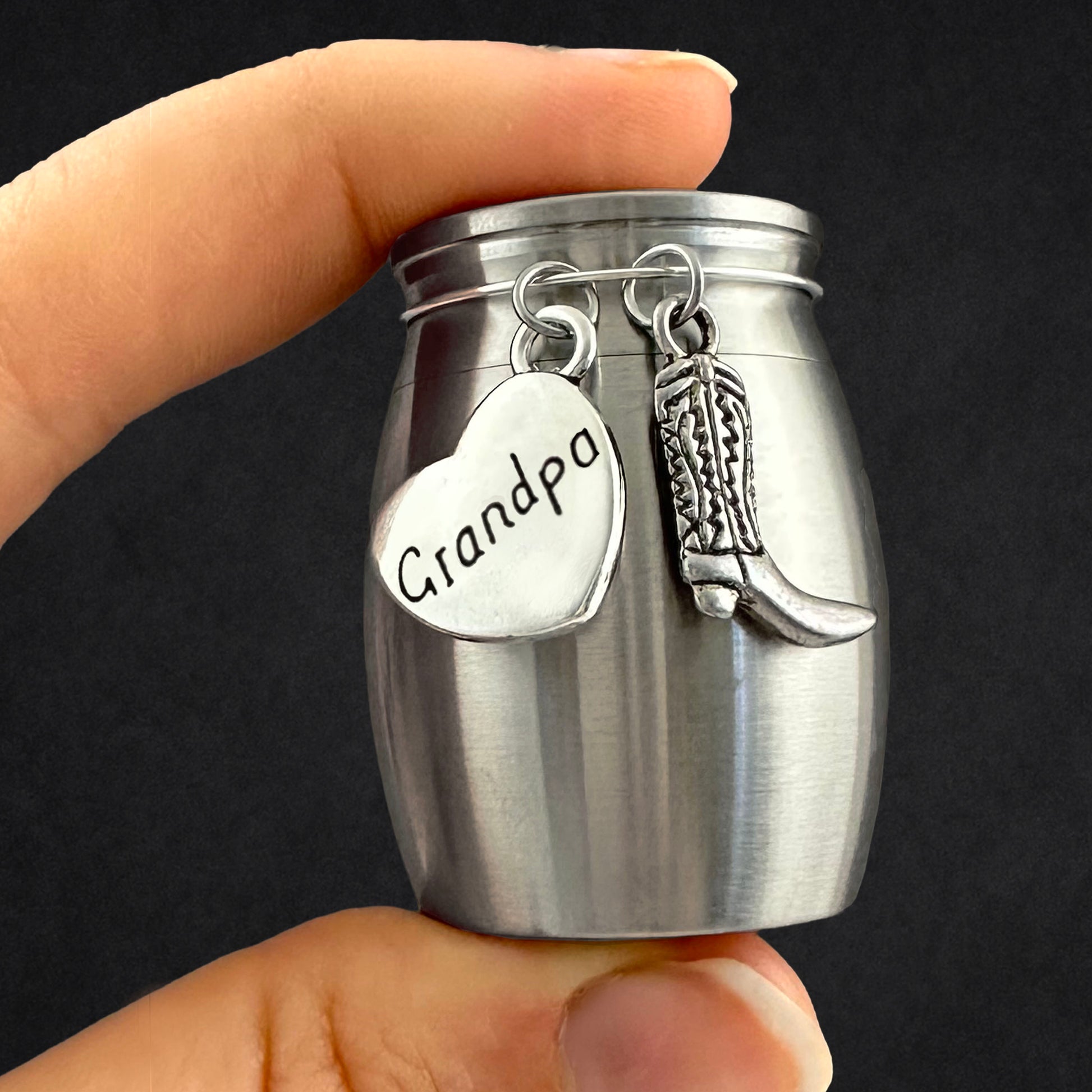 A hand holding a small brushed silver metal keepsake urn with hanging charms, including a heart-shaped tag stamped with the name 'Grandpa' and a cowboy boot charm, against a dark background.