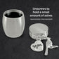 A brushed silver metal cremation urn shown unscrewed and open on a dark background with two charms hanging from the lid: a heart charm engraved with 'Mom' and a cardinal bird charm. White text on the right explains that the urn holds approximately 1 tablespoon of ashes.