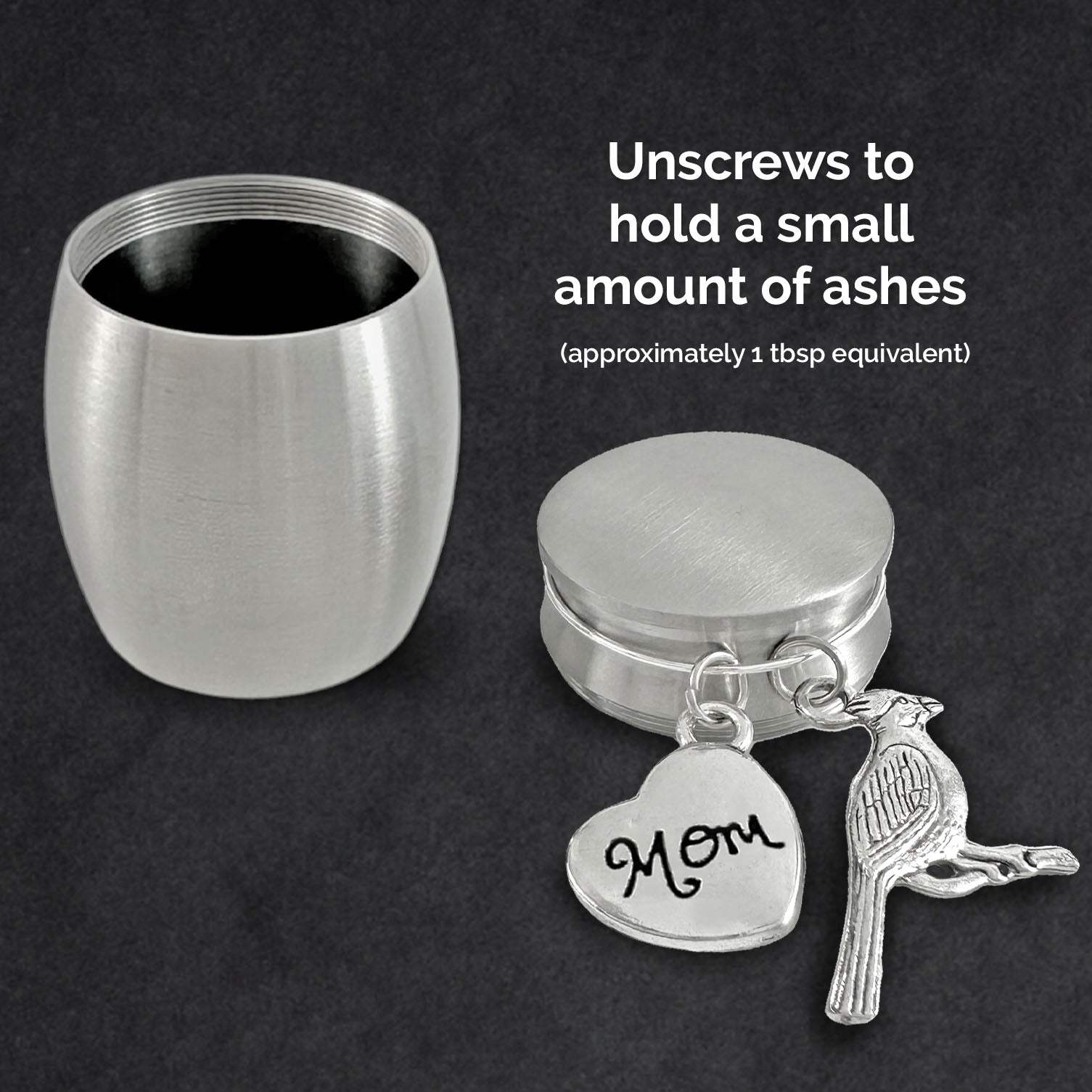 A brushed silver metal cremation urn shown unscrewed and open on a dark background with two charms hanging from the lid: a heart charm engraved with 'Mom' and a cardinal bird charm. White text on the right explains that the urn holds approximately 1 tablespoon of ashes.