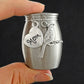 A small brushed silver keepsake urn with a heart-shaped charm engraved 'Mom' and a cardinal bird charm, held by two fingers against a dark background.