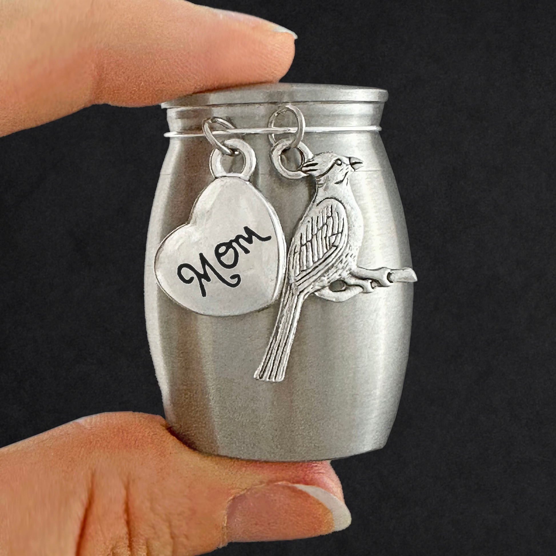 A small brushed silver keepsake urn with a heart-shaped charm engraved 'Mom' and a cardinal bird charm, held by two fingers against a dark background.