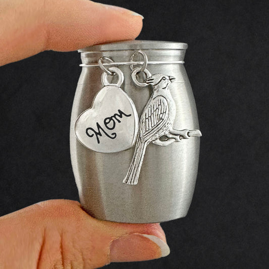 A small brushed silver keepsake urn with a heart-shaped charm engraved 'Mom' and a cardinal bird charm, held by two fingers against a dark background.