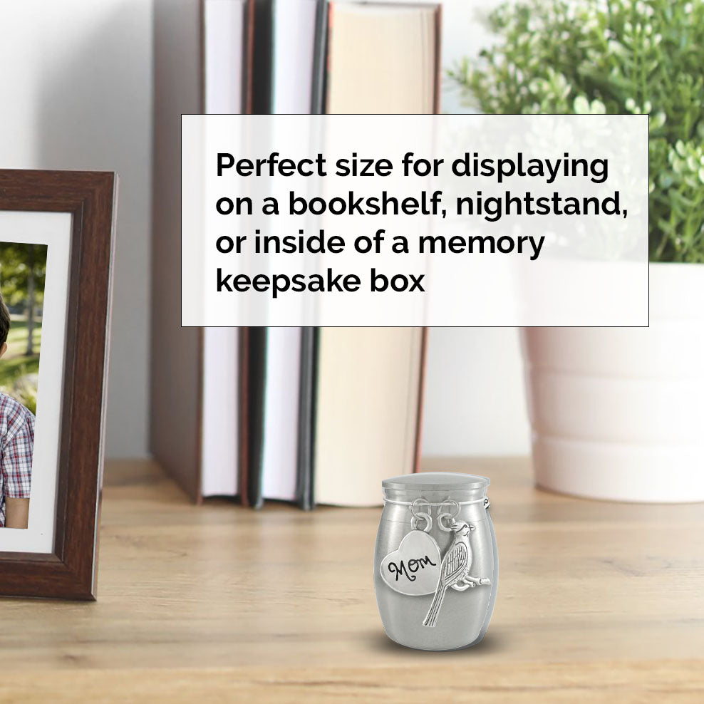 A small silver keepsake urn with a bird and 'Mom' engraving sits on a wooden bookshelf top in a styled home setting, with a text overlay explaining its use for bookshelves, nightstands, or memory keepsake boxes.