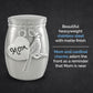 A silver-toned metal jar-shaped keepsake urn with a matte finish, featuring a hanging heart charm engraved with 'Mom' and a cardinal bird charm. The product is shown against a dark textured background with descriptive text on the right. The descriptive text reads: beautiful heavyweight stainless steel with matte finish. Mom and cardinal charms adorn the front as a reminder that mom is near.