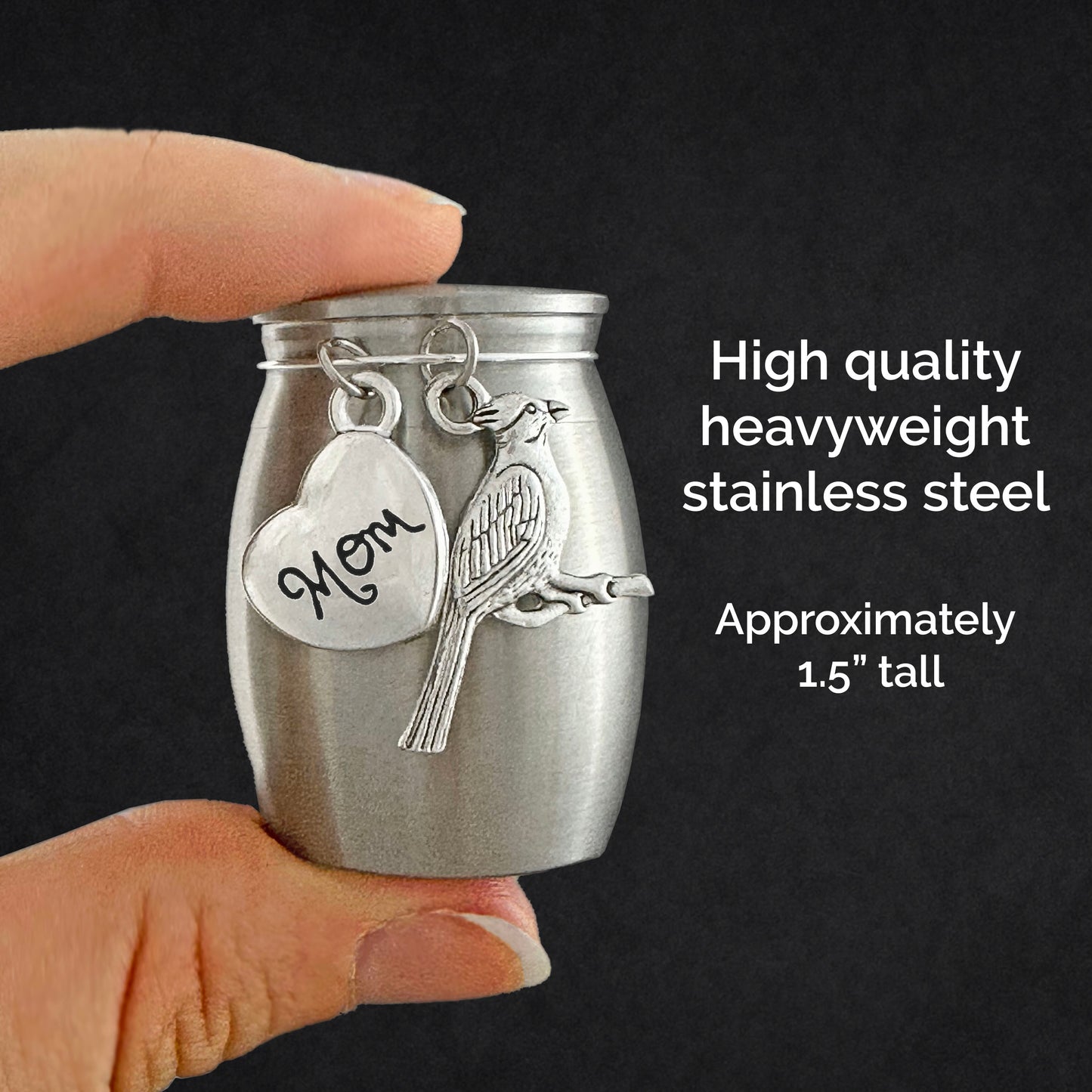 A hand holds a small stainless steel keepsake urn against a dark background, with text on the right reading 'High quality heavyweight stainless steel' and 'Approximately 1.5" tall.