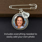 A silver safety pin style keepsake with a round photo frame and a small heart charm engraved 'Mom', shown on a dark wood surface with text noting the photo can be added.