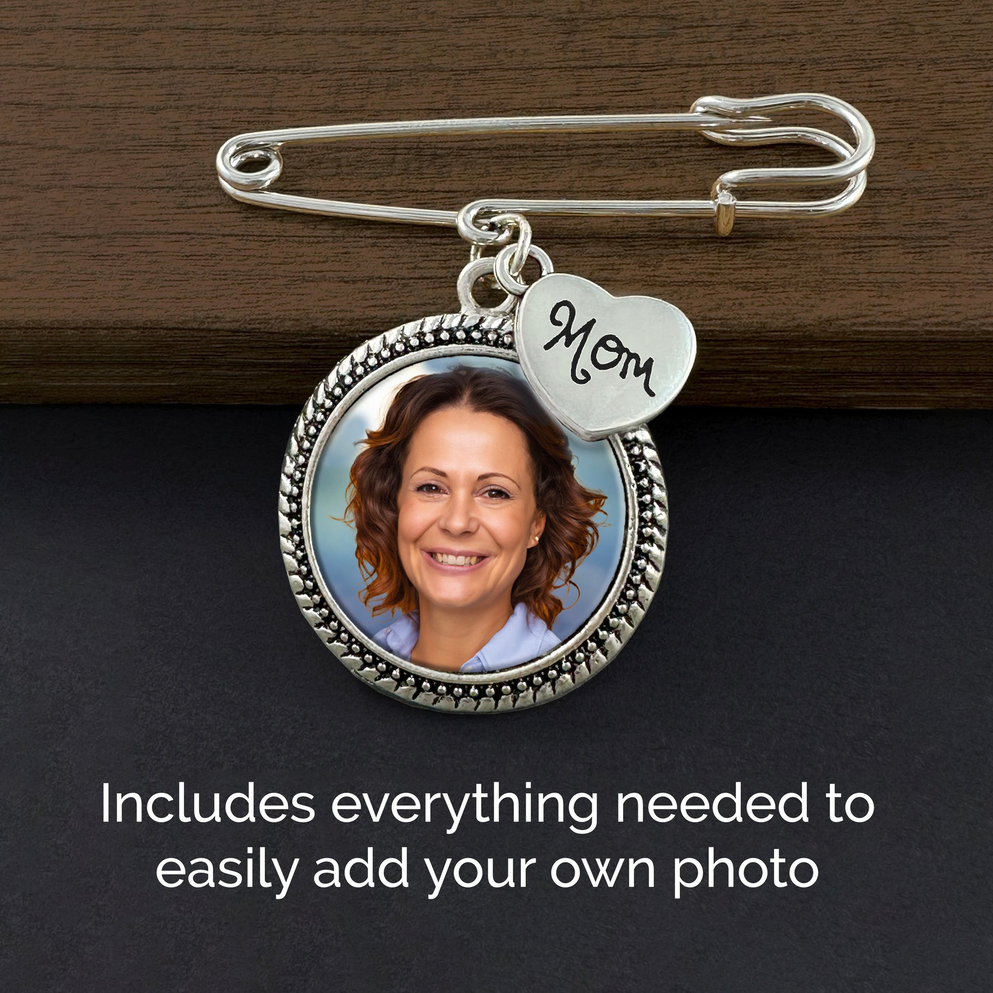 A silver safety pin style keepsake with a round photo frame and a small heart charm engraved 'Mom', shown on a dark wood surface with text noting the photo can be added.