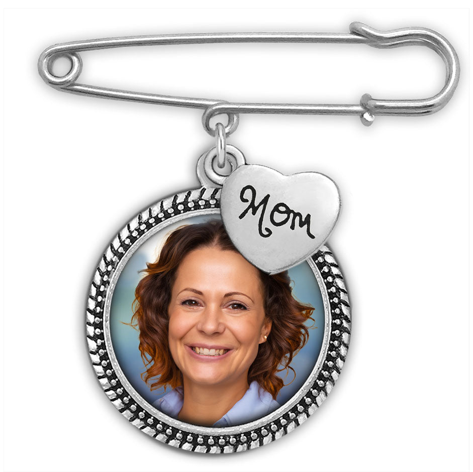 A silver safety pin brooch featuring a round photo frame and a dangling heart charm engraved with 'Mom', shown on a plain white background.