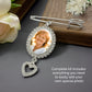 A silvet wedding bouquet photo keepsake pin with an oval rhinestone border holding a portrait sample photo, with a dangling heart-shaped rhinestone charm, styled on a dark surface with flowers and a wooden block. Text on the image reads: 'Complete kit includes everything you need to easily add your own special photo'.