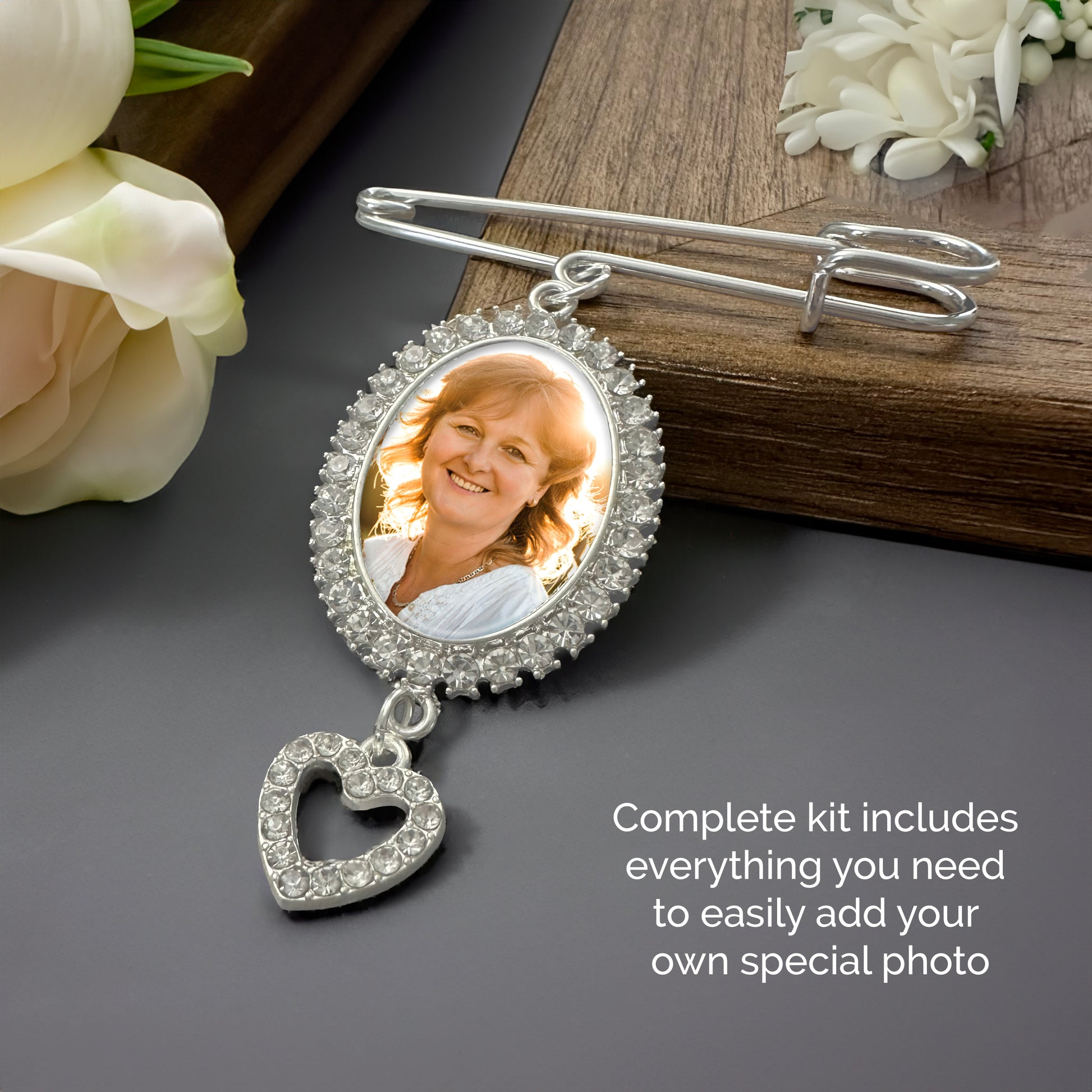 A silvet wedding bouquet photo keepsake pin with an oval rhinestone border holding a portrait sample photo, with a dangling heart-shaped rhinestone charm, styled on a dark surface with flowers and a wooden block. Text on the image reads: 'Complete kit includes everything you need to easily add your own special photo'.