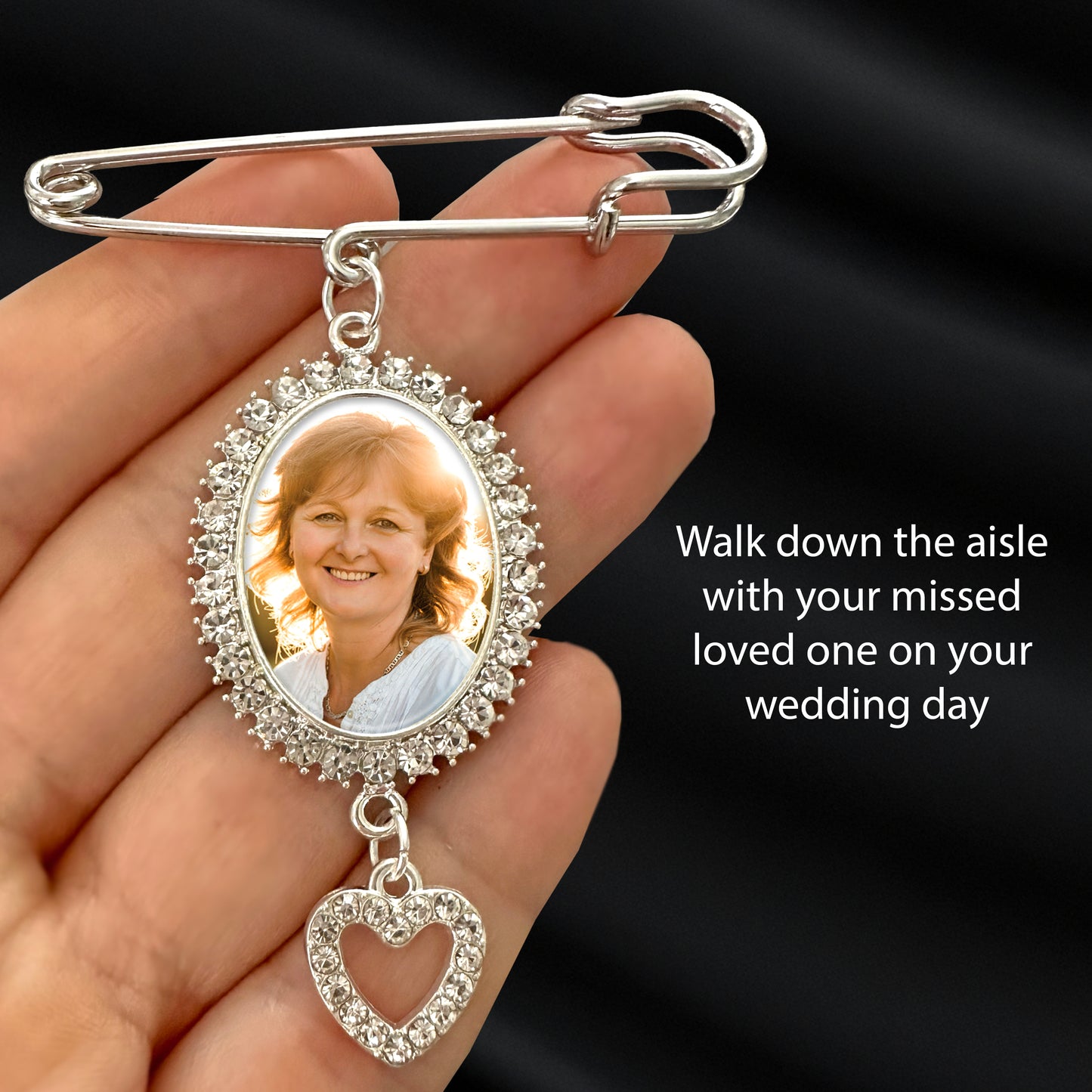 A silver rhinestone photo wedding pin with an oval photo frame and dangling heart charm, held in a hand against a dark satin background. The sample photo insert shows a smiling woman, and the text reads: 'Walk down the aisle with your missed loved one on your wedding day.