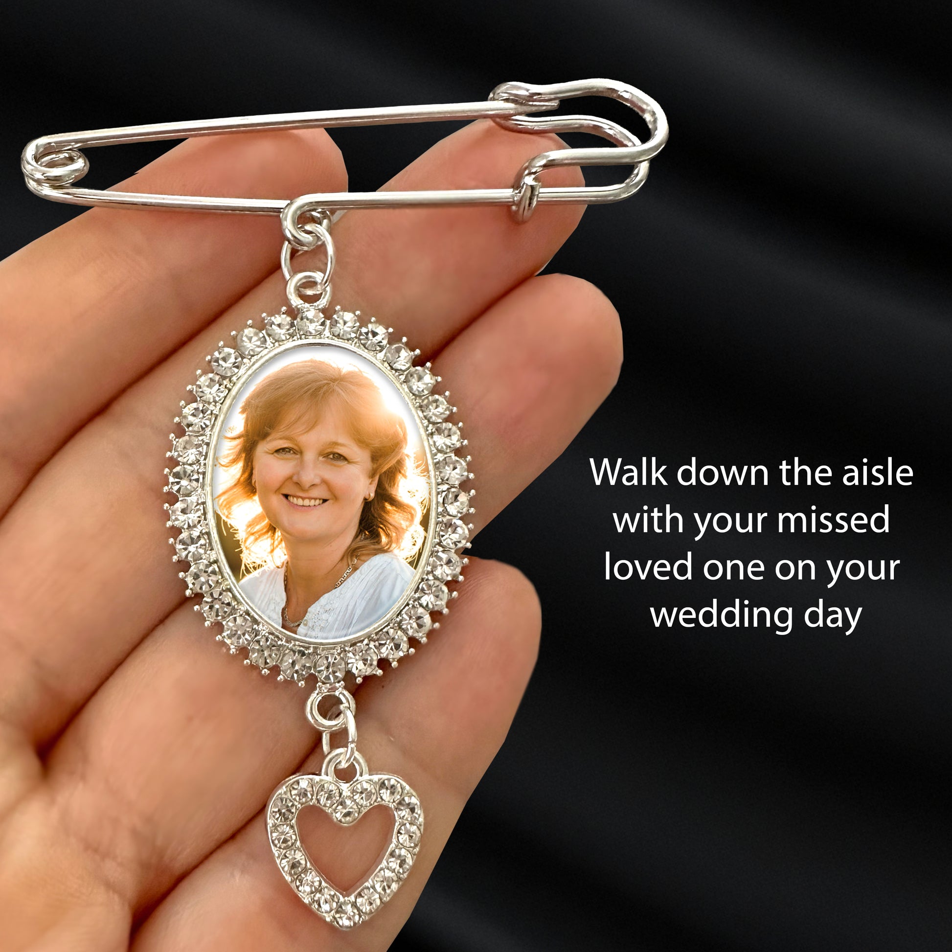 A silver rhinestone photo wedding pin with an oval photo frame and dangling heart charm, held in a hand against a dark satin background. The sample photo insert shows a smiling woman, and the text reads: 'Walk down the aisle with your missed loved one on your wedding day.