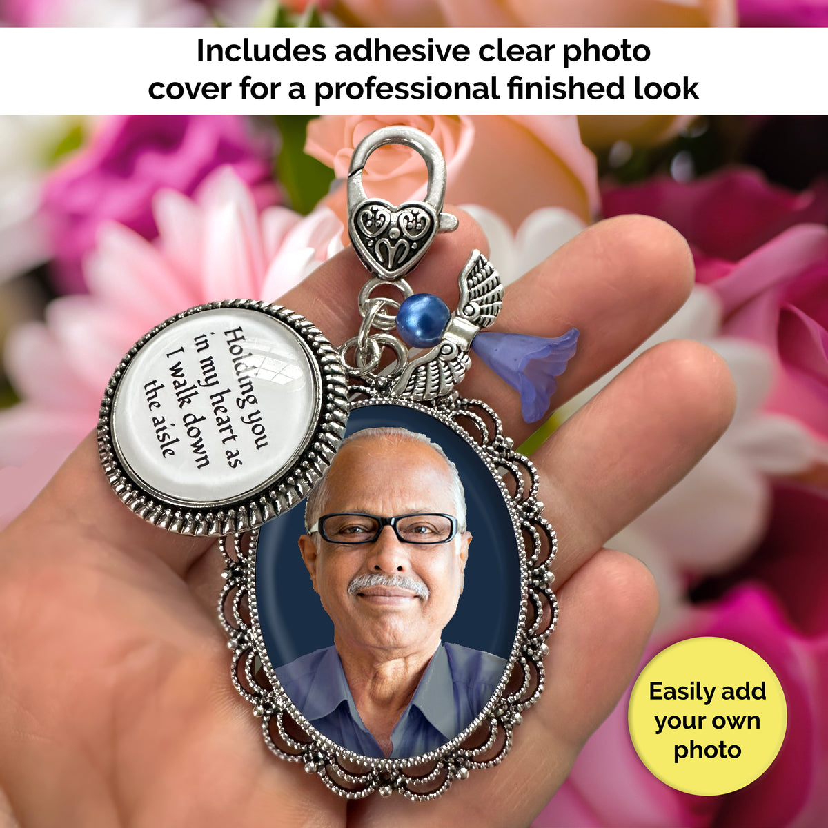 A hand holds a silver-toned memorial pendant keychain with a photo of a man, a round silver charm with text, and decorative blue angel and heart shaped clip. The background is blurred pink flowers, with text overlays reading 'Includes adhesive clear photo cover for a professional finished look' and 'Easily add your own photo'.