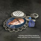 A silver-toned keepsake wedding bouquet photo charm with ornate filigree detailing, shown laying on its side. Has a round photo charm with printed words, and a blue guardian angel  charm and a heart-shaped clasp on a dark textured background.