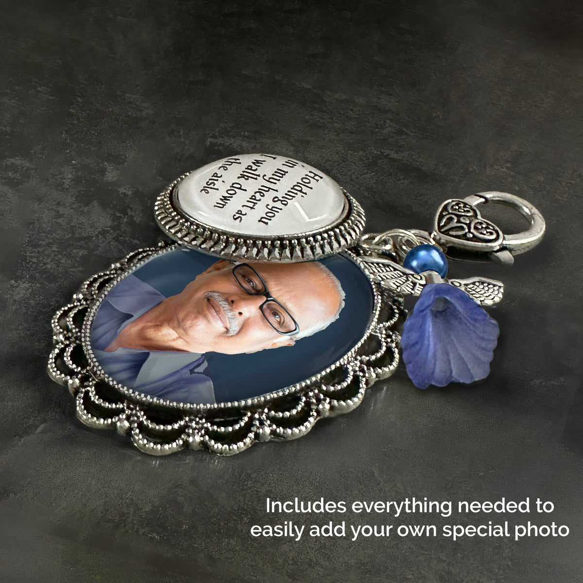 A silver-toned keepsake wedding bouquet photo charm with ornate filigree detailing, shown laying on its side. Has a round photo charm with printed words, and a blue guardian angel  charm and a heart-shaped clasp on a dark textured background.
