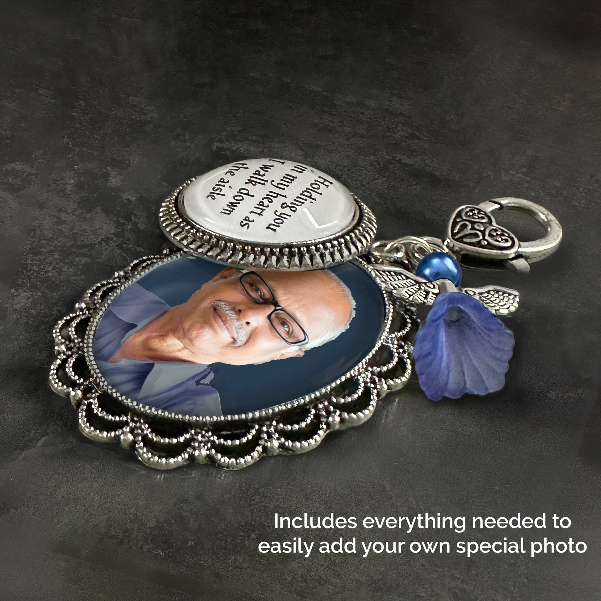 A silver-toned keepsake wedding bouquet photo charm with ornate filigree detailing, shown laying on its side. Has a round photo charm with printed words, and a blue guardian angel  charm and a heart-shaped clasp on a dark textured background.