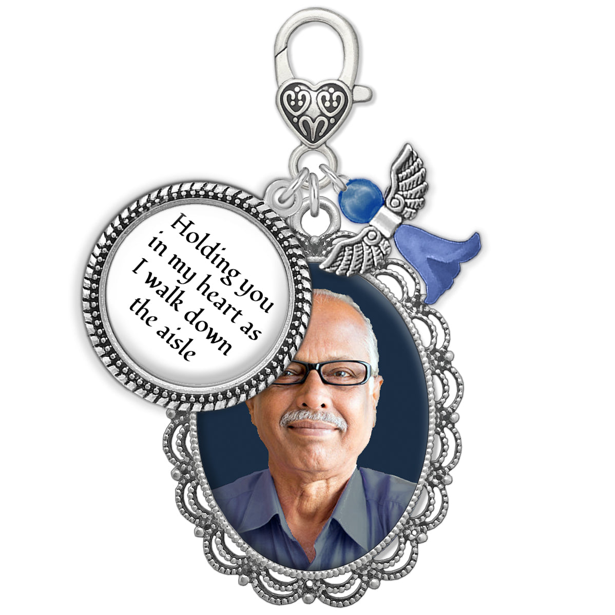 A memorial pendant charm with a silver-toned filigree frame holding a photo of a man, a heart-shaped clip on top of charm with text reading 'Holding you in my heart as I walk down the aisle,' a small blue angel charm, shown on a white background.
