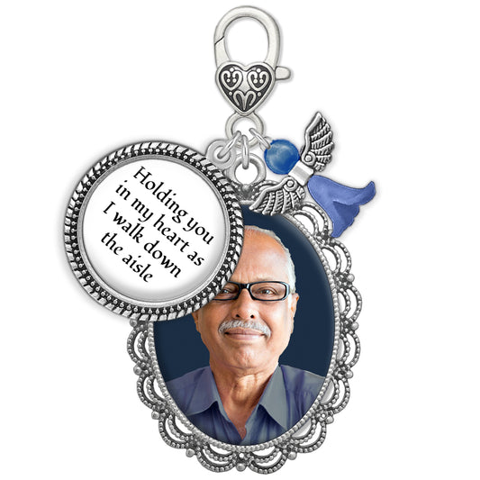 A memorial pendant charm with a silver-toned filigree frame holding a photo of a man, a heart-shaped clip on top of charm with text reading 'Holding you in my heart as I walk down the aisle,' a small blue angel charm, shown on a white background.